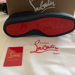Mörkblå sneakers från Christian Louboutin - Snygga mörkblå sneakers från Christian Louboutin med den ikoniska röda sulan. Skorna har snörning och en stilren design som passar perfekt för en trendig look. Tillverkade i Italien och levereras med originalförpackning och dustbag.