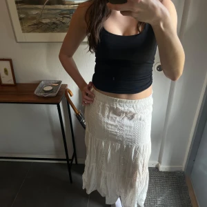 Vit långkjol med spetsdetaljer - Säljer en vacker vit långkjol med spetsdetaljer. Kjolen har en elastisk midja för bekväm passform och ett romantiskt utseende med volanger. Perfekt för sommarens varma dagar. Står inte storlek i kjolen men skulle säga en S. Måtten är 80cm i längden och 33 i midjan 