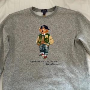 Grå tröja med Polo Bear från Ralph Lauren storlek L i barn - Säljer en grå tröja från Ralph Lauren med det ikoniska Polo Bear-motivet på framsidan. Tröjan har långa ärmar och är perfekt för en avslappnad stil. Den är tillverkad i ett mjukt material för extra komfort.