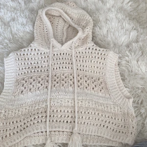 Beige stickad väst med huva från Zara - Säljer en beige stickad väst från Zara med huva och dekorativa tofsar. Västen har ett fint hålmönster och är perfekt för lager-på-lager-stil. Den är ärmlös och har en avslappnad passform.