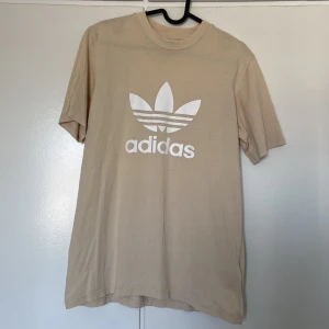 Beige t-shirt från Adidas - Säljer en beige t-shirt från Adidas med det klassiska trefoil-logotypen i vitt på framsidan. T-shirten har en lös passform och är kortärmad, perfekt för en avslappnad stil.