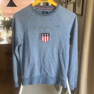 Blå tröja från GANT - Säljer en snygg blå tröja från GANT med ett broderat emblem på bröstet. Tröjan har långa ärmar och en rund halsringning. Perfekt för en avslappnad stil.