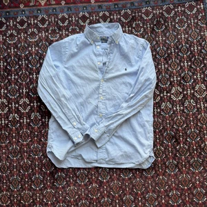 Ljusblå skjorta från Polo Ralph Lauren - Säljer en stilren ljusblå skjorta från Polo Ralph Lauren. Skjortan har en klassisk krage och knappar framtill. Den är slim fit och har ett broderat logotypmärke på bröstet. Perfekt för en snygg och avslappnad look.