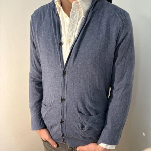 Blå kofta från Guess - Säljer en stilren blå kofta från Guess med knappar framtill och praktiska fickor. Koftan har en huva och långa ärmar, perfekt för en avslappnad look. Passar bra till både jeans och chinos. Bra skick, inga defekter. Modellen är 182cm/67kg