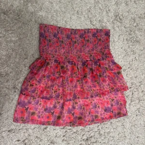 Blommig kortkjol med volang - Söt blommig kortkjol i rosa och lila toner med volangdetaljer. Perfekt för sommarens varma dagar. Den har en elastisk midja för bekväm passform. Helt oanvänd och säljs inte längre.