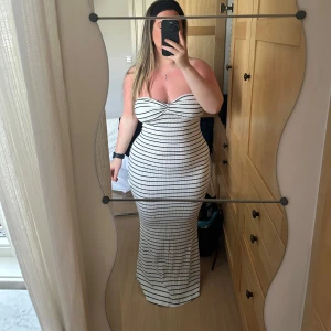  klänning - Snygg axelbandslös långklänning i vit med svarta ränder. Perfekt för en stilren look med en tight passform som framhäver figuren. Klänningen är enkel men elegant och passar bra till olika tillfällen.