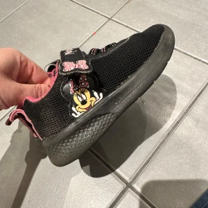 Svarta och rosa sneakers med Minnie Mouse - Söta svarta sneakers med rosa detaljer och Minnie Mouse-motiv. Skorna har kardborreband med en rosett och texten 'Minnie'. Perfekta för barn som älskar Disney!