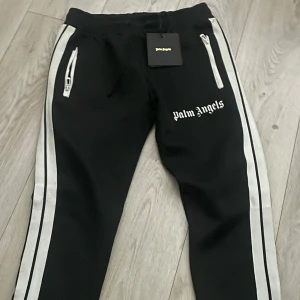Svarta Palm Angels byxor - Snygga svarta Palm Angels byxor med vita sidoremsor och dragkedjor på fickorna. Byxorna har en elastisk midja och Palm Angels-loggan tryckt på benet. Perfekta för en avslappnad stil. Säljer billigt bra för eget bruk men även resella. 