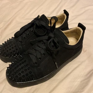 Christian Louboutin  -  Är mest intresserad av byten Snygga svarta sneakers med nitar på tån och sidorna. Skorna har en klassisk snörning och en iögonfallande röd sula. Perfekta för att ge din outfit en edgy touch. Passar 39 till 40/41