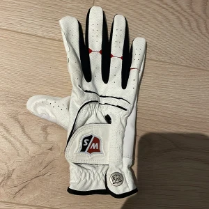 Vit golfhandske från Wilson Staff - Säljer en vit golfhandske till höger hand från Wilson Staff med svarta och röda detaljer. Handsken har perforeringar för bättre ventilation och en justerbar kardborrestängning för optimal passform. Perfekt för golfentusiaster som vill ha stil och funktionalitet på banan. 🏌️‍♂️