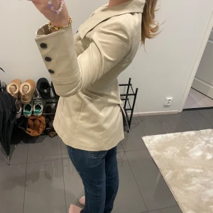 Beige kappa med knappar - Snygg beige kappa med stora knappar framtill och en klassisk krage. Jackan har en figursydd passform och långa ärmar. Perfekt för en stilren look.