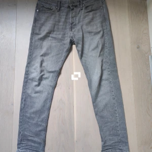 Grå jeans från Jack & Jones - Snygga grå jeans från Jack & Jones med klassisk femficksdesign. De har en knappgylf och bälteshällor, vilket gör dem perfekta för en avslappnad stil. Passformen är modern och de är tillverkade i ett slitstarkt material.