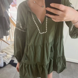 Grön klänning med broderi - Säljer en söt grön klänning med v-ringning och broderade detaljer i vitt. Klänningen har långa ärmar och en luftig passform med volanger. Perfekt för en avslappnad stil.