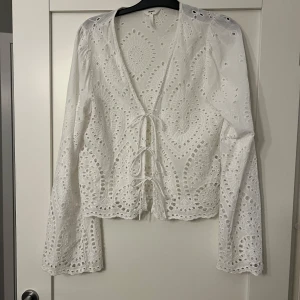 Vit blus från Object - Säljer en vit blus från Object med vackert broderat mönster och knytning framtill. Blusen har långa ärmar och en v-ringad design som ger en elegant touch. Perfekt för både vardag och speciella tillfällen.