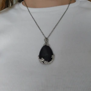 Lila droppformat halsband med silverdetaljer - Elegant halsband med en lila droppformad kristall infattad i silver. Hänget har vackra blommönster och en kedja i silverfärg. Perfekt för att ge en färgklick till din outfit.