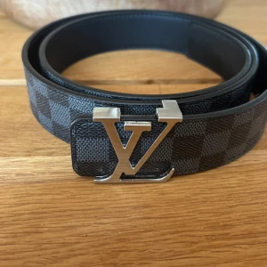 Louis Vuitton rutig svart skinnbälte - Säljer ett Louis Vuitton bälte i svart skinn med klassiskt rutigt mönster och en stor silverfärgad LV-logga som spänne. Bältet har en stilren och lyxig känsla, perfekt för dig som vill sticka ut med en ikonisk accessoar från ett av världens mest kända modehus. Priset är förhandlingsbart 