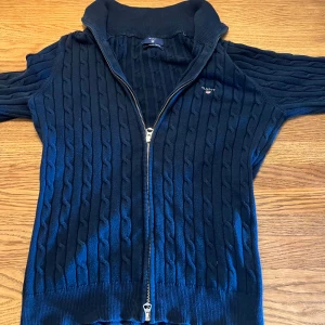 Marinblå kabelstickad kofta från Gant - Kabelstickad marinblå kofta från Gant med hel dragkedja och hög krage. Klassisk design med ribbade muddar och diskret logga på bröstet. Perfekt för lager-på-lager och snygg till jeans eller chinos.