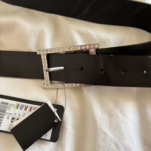 Svart bälte med glittrigt spänne New Yorker - Snyggt svart bälte från New Yorker med ett fyrkantigt spänne dekorerat med glittrande stenar. Bältet är i slätt material och har flera hål för justerbar passform. Perfekt accessoar för att lyfta din outfit med lite extra bling.