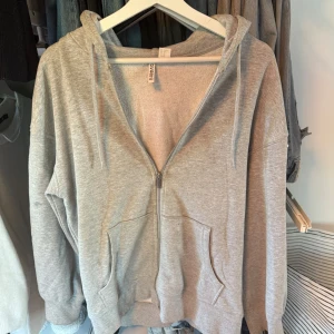 Grå hoodie med dragkedja från H&M - En klassisk grå hoodie från H&M med dragkedja framtill, huva med snörning och två stora fickor. Mjuk bomullsblandning och långärmad modell, perfekt för chill dagar eller när du vill ha en enkel och clean look. På bild två så ser man att den är en liten fläck som tyvärr inte går bort... annars är den fin i skick! 