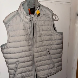 Ljusgrå dunväst från Parajumpers - Säljer en ljusgrå dunväst från Parajumpers med quiltad design och hög krage. Västen har dragkedja framtill, två snedställda fickor och en gul detalj vid halsen. Perfekt för lager-på-lager och snygg till både hoodie och t-shirt.
