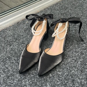 Svarta pumps med pärldetaljer och rosett - Eleganta svarta pumps i satin med spetsig tå, låg klack och dekorativa pärlor runt vristen. Skorna har en bred rosett i satin som knyts runt ankeln för extra statement. Perfekta för dig som vill sticka ut med en lyxig och trendig look.