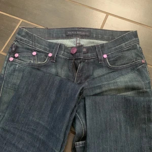 Rock & Republic jeans med lila detaljer - Mörkblå jeans från Rock & Republic med snygga lila och rosa detaljer, inklusive broderade bakfickor och lila knappar. Klassisk femficksmodell i jeansmaterial med raka ben och cool faded effekt. Perfekta för dig som gillar statement-jeans med unik touch. Jeansen har små slitage som visas på bilden. 