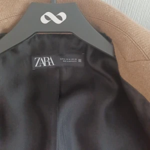 Beige kappa från Zara i ullmix - Snygg beige kappa från Zara i klassisk modell med dubbelknäppning och bred krage. Jackan har raka fickor, knappar vid ärmslut och är tillverkad i en ullmix som ger en lyxig känsla. Perfekt för dig som vill ha en stilren och tidlös look.
