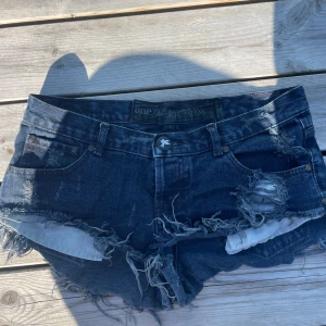 Mörkblå slitna jeansshorts One Teaspoon - Riktigt snygga mörkblå lågmidjade jeansshorts från One Teaspoon, fransiga kanter och slitna detaljer både fram och bak. Klassisk femficksmodell med låg midja och coolt, avslappnat vibe. Perfekta för sommardagar när du vill sticka ut lite extra. Skulle säga att de passar S/M