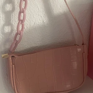 Rosa axelväska med kedjedetalj - Säljer en ljusrosa axelväska i syntetmaterial med croco-mönster. Väskan har en chunky plastkedja som detalj på axelremmen och stängs med dragkedja. Guldiga metalldetaljer och insida i matchande rosa tyg. Perfekt för dig som gillar trendiga accessoarer.