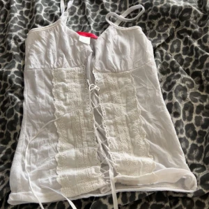 Vit korsett-topp med snörning - Supersöt vit korsett-topp med tunna axelband och snygg snörning framtill. Toppen har spetsdetaljer och broderade partier längs framsidan som ger en romantisk vibe. Perfekt för dig som gillar en feminin och trendig look.