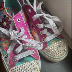 Twinkle Toes sneakers med glitter och blommor - Färgglada Twinkle Toes sneakers från Skechers med blommigt mönster, glittriga rosa och silverdetaljer samt strass på tån. Skorna har grå snörning och en platt sula. Perfekta för dig som gillar att sticka ut med coola detaljer och mycket färg.