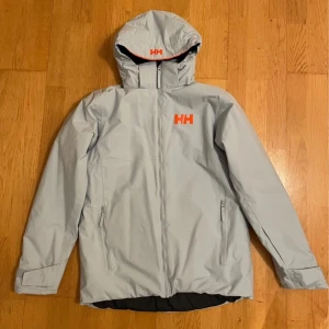 Ljusblå skidjacka från Helly Hansen - Snygg ljusblå skidjacka från Helly Hansen med orange logga på bröstet och ryggen. Jackan har huva, hel dragkedja framtill och två fickor med dragkedja. Insidan är svart och har snölås. Är stl 176 men motsvarar xs-m. Endast använd en skidsäsong. Ny pris är ca 2000