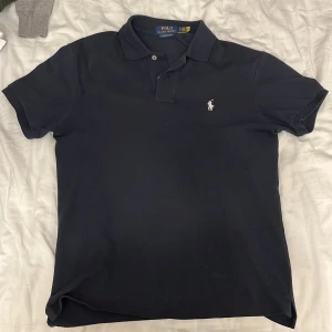 Marinblå pikétröja från Polo Ralph Lauren - Klassisk marinblå pikétröja från Polo Ralph Lauren med vit broderad logga på bröstet. Tröjan har korta ärmar, krage och två knappar framtill. Tillverkad i mjuk bomull som känns skön mot huden. Perfekt för en clean och stilren look.