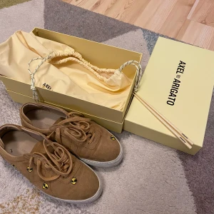 Axel Arigato sneakers i original förpackning  - Axel arigato sneakers beige/bruna med aliens på. Original förpackning följer med som inkluderar en gym påse som man kan ha skorna i eller använda för annat. Dessutom ingår matpinnar.   Storlek: 42 (se bild) Skick: använt några gånger vilket syns på bilderna, men inte så mycket. De har fräsch form kvar och de smuts som syns på sulan går att tvätta bort.