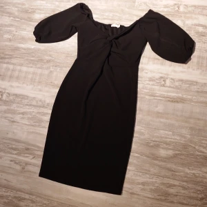 Svart Belle Sleeve klänning NLY Trend (oanvänd) - Svart klänning från NLY Trend med snygg v-ringning och puffiga trekvartsärmar. Modellen är figurnära med knutdetalj framtill och går till knäna. Perfekt för dig som gillar stilren och trendig look.