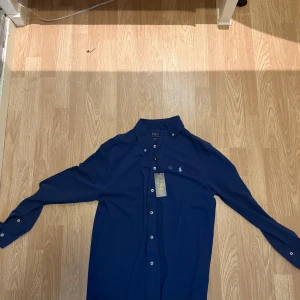 Marinblå skjorta från Polo Ralph Lauren - Snygg marinblå skjorta från Polo Ralph Lauren med klassisk krage och vita knappar. Skjortan har lång ärm och ikoniska broderade loggan på bröstet. Tillverkad i mjuk bomull som känns skön mot huden. Perfekt för dig som gillar stilrena och tidlösa plagg.