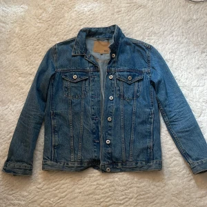 Klassisk blå jeansjacka från 157 XS - Snygg blå jeansjacka från 157 i klassisk modell med raka linjer, bröstfickor med knappar och silverfärgade detaljer. Jackan är tillverkad i jeansmaterial och har lång ärm. Perfekt för dig som gillar en tidlös streetstil.