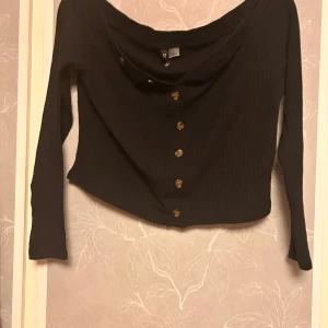 Svart ribbad topp från Divided - Svart långärmad topp från Divided med ribbad struktur och snygg offshoulder-ringning. Toppen är croppad och har dekorativa knappar framtill som ger en extra touch. Perfekt för dig som gillar en stilren men trendig look.