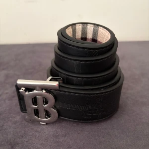 Svart Burberry bälte med B-spänne - Stilrent svart bälte från Burberry i skinn med klassiskt rutmönster och silverfärgat B-formad metallspänne. Insidan har den ikoniska Burberry-rutiga designen i beige, svart och vinrött. Perfekt accessoar för att lyfta din outfit.