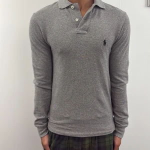 Grå långärmad pikétröja Polo Ralph Lauren - Snygg grå långärmad pikétröja från Polo Ralph Lauren i slim fit. Tröjan har klassisk krage, knäppning med två knappar och den ikoniska broderade loggan på bröstet. Perfekt för en clean och stilren look.