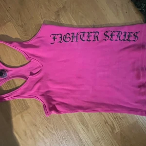Y2k rosa linne - Cool rosa linnetopp från Fighter Series med svart tryck framtill och bak, där det står 'Fighter Series' och en stor dödskalle med vingar. Modellen är ärmlös med brottarrygg och ribbad struktur. Perfekt för dig som gillar edgy och sportig stil.