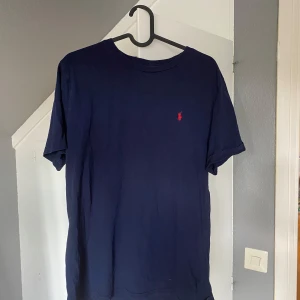 Mörkblå t-shirt från Polo Ralph Lauren - Snygg mörkblå t-shirt från Polo Ralph Lauren i bomull. Klassisk passform med korta ärmar och rund hals. Liten röd broderad logga på bröstet som ger en clean och stilren look. Det är xl barn och det motsvarar som storlek S