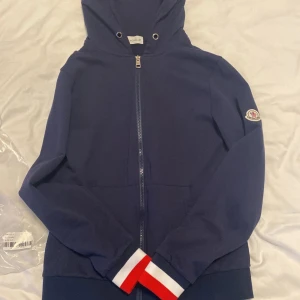 Marinblå Moncler hoodie med dragkedja - Snygg marinblå hoodie från Moncler med hel dragkedja och dubbla zippar. Huva med röda och vita ränder på insidan, samt klassisk Moncler-logga på ärmen. Pris går att diskutera. De står storlek M men passar inte M passar xs och S. Använt den 3 gånger 