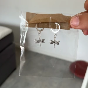 Silvriga örhängen med trollslända - Snygga silvriga örhängen med hängande trollsländor dekorerade med små glittriga stenar. Perfekta för att ge din look en cool och unik touch. Lätta att bära och passar till många olika stilar.