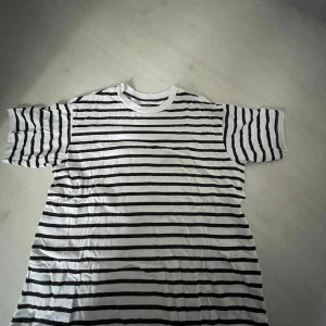 Randig t-shirt från 157, storlek S - Vit t-shirt med svarta horisontella ränder från 157. Klassisk rund halsringning och korta ärmar. Tillverkad i mjuk bomull, perfekt för en chill och stilren look.