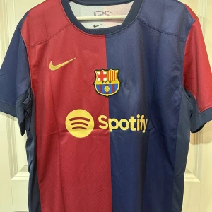 FC Barcelona Lamine Yamal matchtröja - Snygg FC Barcelona matchtröja från Nike med Lamine Yamal och nummer 19 på ryggen. Tröjan är kortärmad och har klassisk halv-halv design i rött och mörkblått, med klubbmärke, Nike-logga och Spotify som sponsor framtill. Tillverkad i lätt och ventilerande material.