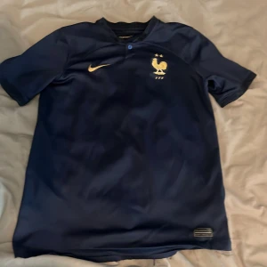 Frankrike Nike fotbollströja marinblå - Säljer en officiell Frankrike fotbollströja från Nike i marinblått med guldiga detaljer. Tröjan har korta ärmar, broderad Nike-logga och FFF-emblem med tupp och två stjärnor. Tillverkad i lätt och ventilerande funktionsmaterial, perfekt för match eller träning.