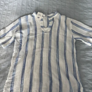 Randig vit och blå henley från Boomil - Snygg henley-skjorta från Boomil i vit och blå med vertikala ränder. Skjortan har korta ärmar, v-ringad hals och är tillverkad i ett luftigt bomullstyg, perfekt för varma dagar. Grekisk design med avslappnad passform och klassisk sommarkänsla.