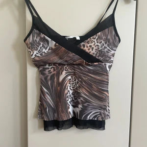 Leopardmönstrad linne med meshdetalj - Säljer ett linne med leopardmönster i bruna, svarta och beige toner. Toppen har smala axelband, omlott-effekt framtill och svart meshdetalj vid bysten samt längst ner. Snyggt flowy material och figurnära passform.