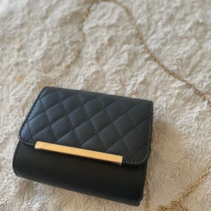 Svart quiltad axelväska med guldkedja - Snygg svart axelväska med quiltat mönster och guldfärgad kedja. Väskan har en stilren gulddetalj framtill och ett fack med dragkedja inuti. Perfekt storlek för det viktigaste och ger en lyxig känsla till din outfit.